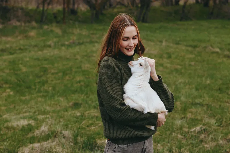 A woman in a dark sweater holds a goat kid in a green pasture, smiling softly. A lifestyle moment with animal affection, care, and companionship in a rural countryside setting.