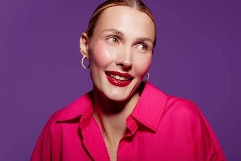 Smiling woman with red lipstick and pink shirt looks away on purple background. Portrait of happy female model with hoop earrings and natural makeup in studio lighting.