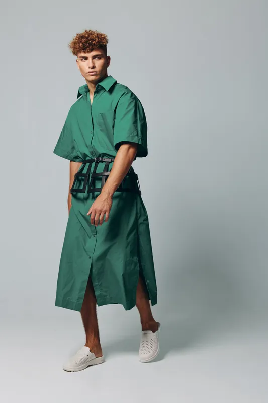 Young man with curly hair wearing green oversized shirt dress and white slip-on shoes posing in studio with neutral background. Fashion model with casual streetwear and modern style.