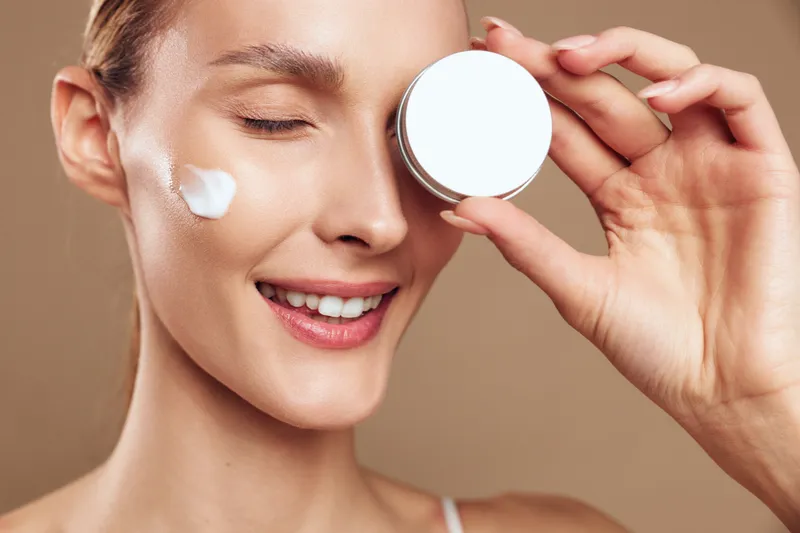 A close-up of a smiling woman applying facial cream in a bright studio, illustrating a skincare routine, hydration, and a healthy glow through moisturizer application and self care.