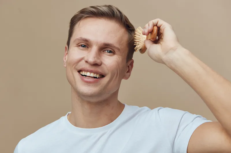 Smiling man using a brush to groom his hair, casual light blue t-shirt, cheerful expression, indoor background, lifestyle moment, male grooming, everyday routine, positive mood, natural lighting, port