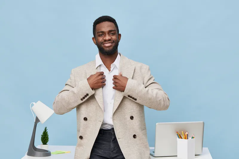 Confident young African American man in stylish beige blazer smiling, adjusting collar, in modern office with laptop, desk lamp, and stationery, relaxed and professional, studio shot, bright backgroun