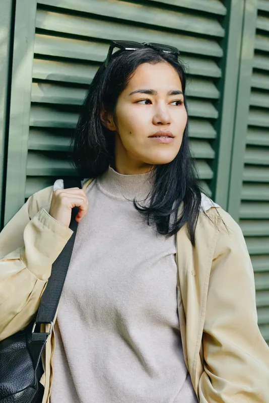 Woman wearing a light beige jacket and gray knit sweater stands beside green shutters, conveying casual street style and thoughtful mood for fashion, urban life, and everyday wear, ready for