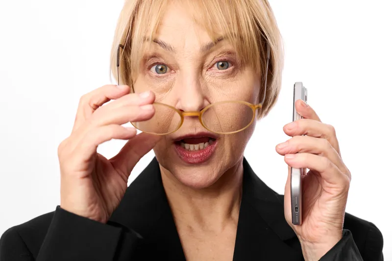 Overwhelmed senior woman in call center headset holding smartphone and adjusting round glasses, anxious surprised look, isolated on white background, mental stress concept