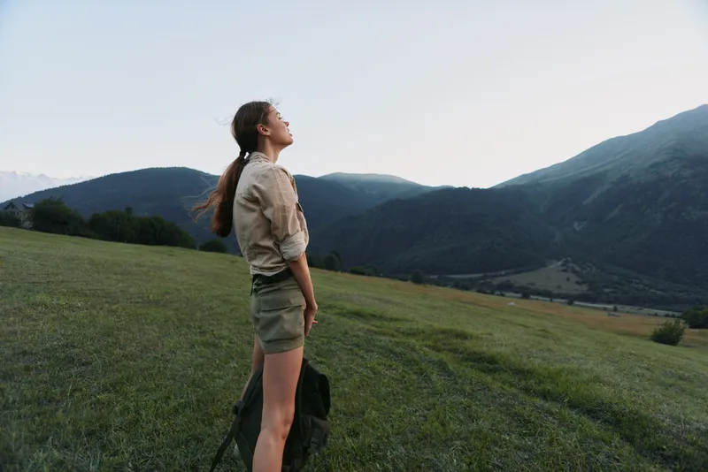 Cheerful woman outdoors, standing in a sunlit meadow with distant mountains, wearing casual shorts and shirt, enjoying fresh air, a peaceful moment, and a relaxed lifestyle