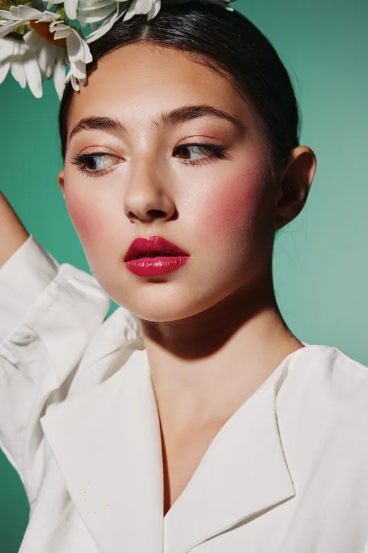 Young woman with red lipstick and natural blush posing in white blouse against green background. Beauty portrait with creative fashion concept and elegant flower detail in hair.