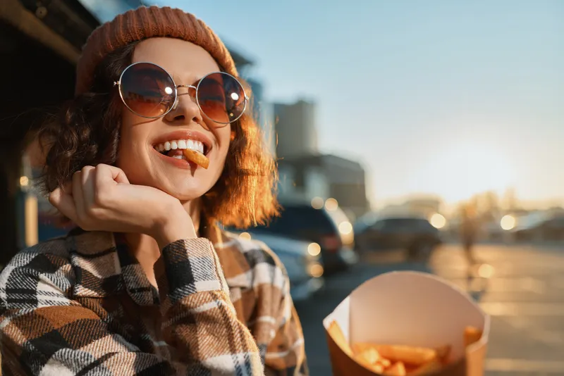 Woman with fries and smile wearing sunglasses and beanie in urban parking at golden hour, candid moment capturing authenticity, candid lifestyle, golden hour glow and emotional storytelling.