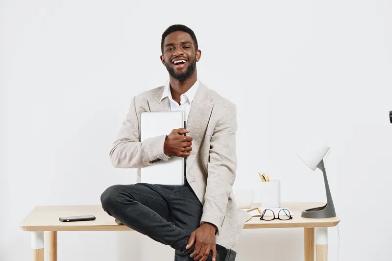 Smiling man in business casual attire holding tablet, sitting on wooden desk with workspace items like glasses, lamp, and notepad, modern office setting, positive professional vibe, bright background,
