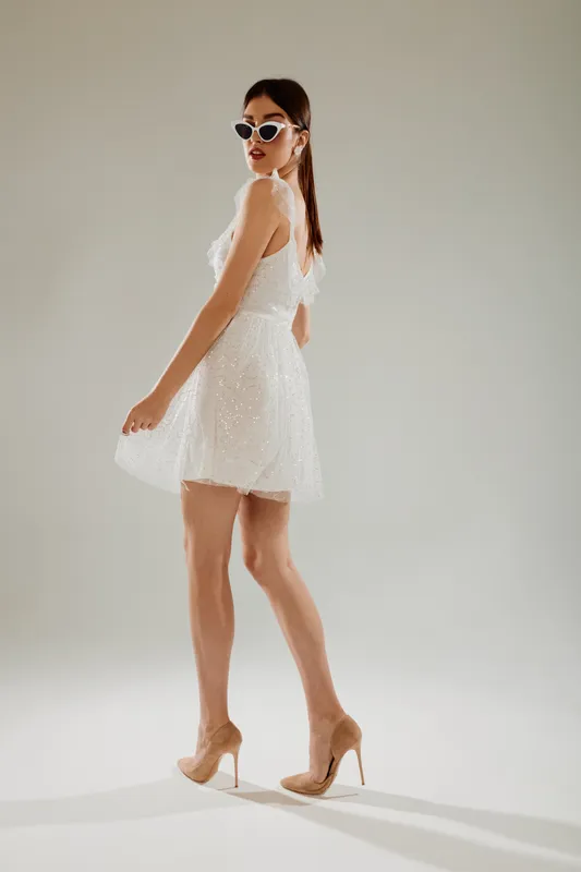 Slim woman in a white dress and sunglasses poses confidently on a light background. Concept of woman beauty fashion with elegance and modern style in studio lighting.