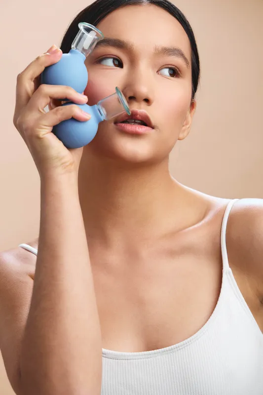 Asian woman in a white top holds a blue perfume bottle close to her face, delivering a soft, confident look in a clean, studio portrait.