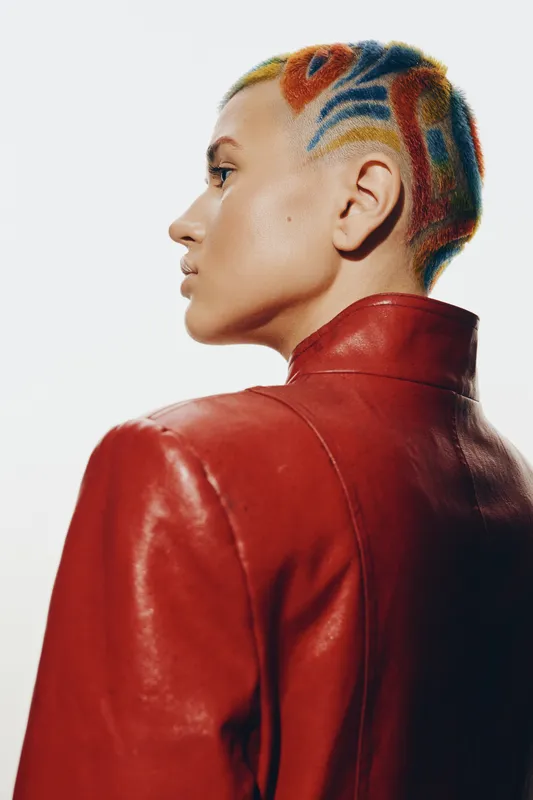 Young woman with colorful short hair and shaved patterns wearing a bold red leather jacket, showcasing modern fashion and edgy urban style in studio lighting.
