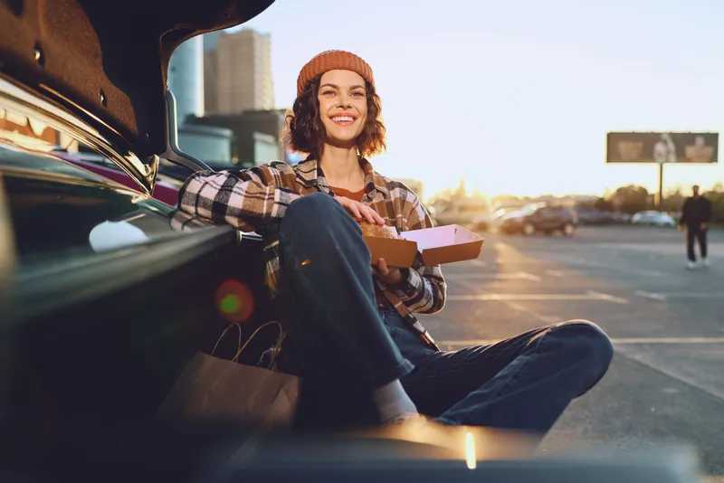 Woman in car trunk eating pizza in a sunny parking lot, smile and relaxed pose, urban golden hour glow, authenticity and candid lifestyle moment with mindful living and emotional storytelling.