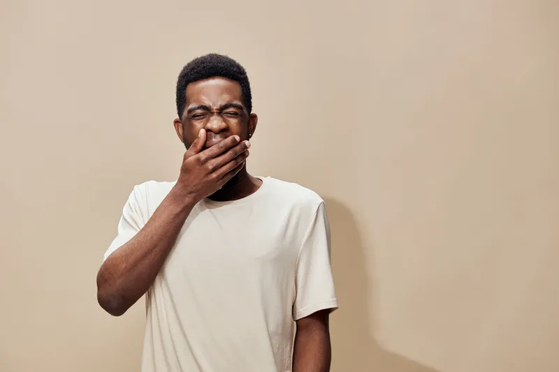 Young African man yawning with a funny expression, wearing a casual cream t shirt against a beige background, conveying tiredness and relaxation
