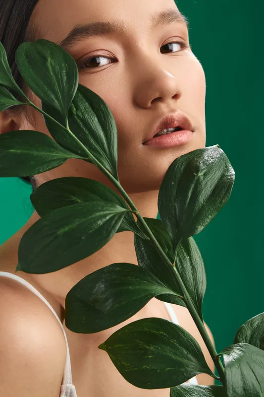 Asian woman is photographed in a stylish studio portrait with lush greenery framing her face, conveying natural beauty, calmness, and graceful presence.