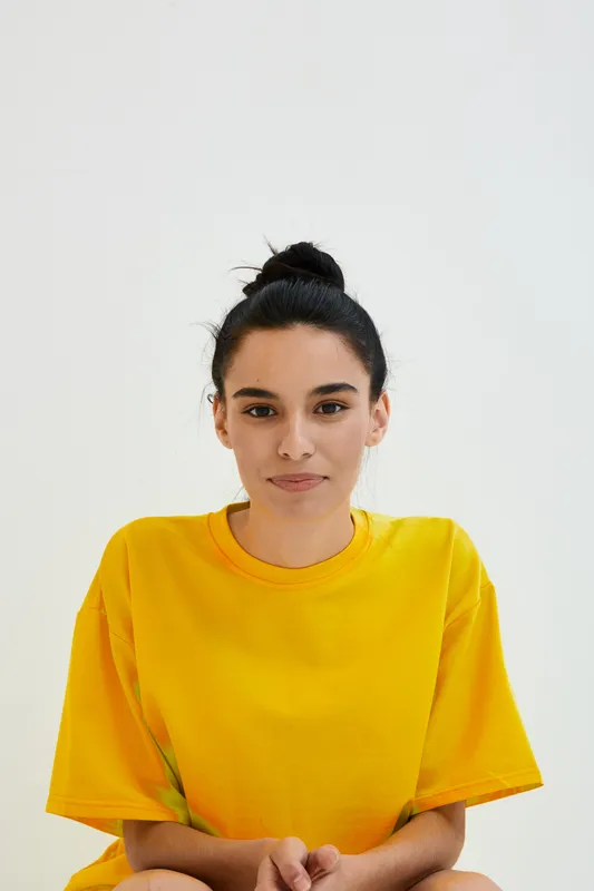 young woman in yellow shirt with dark hair tied up, sitting calmly against plain white background, natural look with minimal makeup and relaxed posture