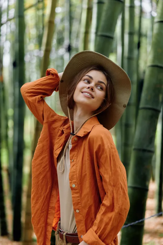 Woman in hat and orange jacket standing among bamboo forest, smiling portrait with relaxed pose, natural light and stylish travel outfit for outdoor lifestyle and nature exploration
