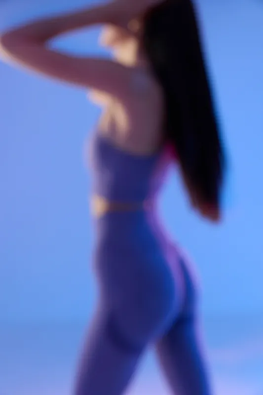 Sport fitness woman in purple activewear posing with arms raised, showing toned body and long dark hair against soft blue background, highlighting health and strength.