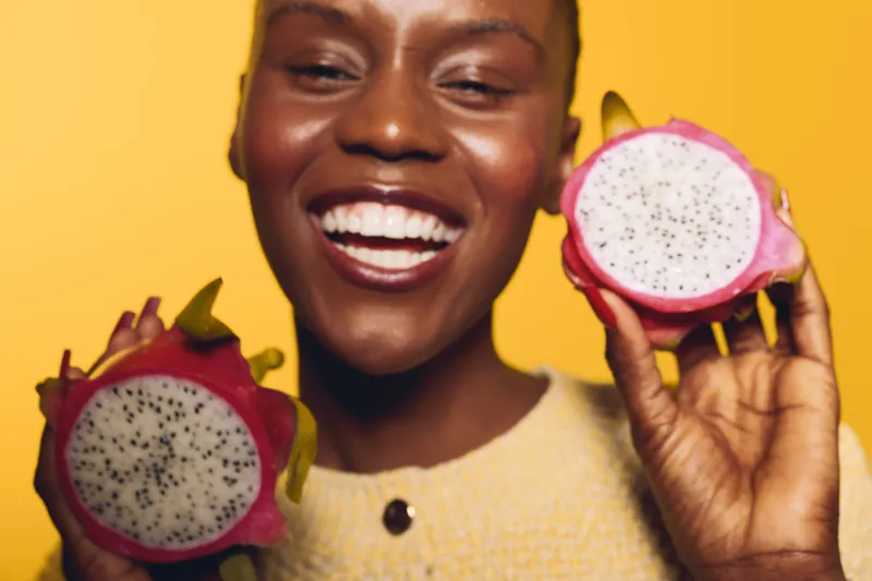 Happy African woman with dark skin holds two halves of dragon fruit, smiling widely against a yellow background. Concept for healthy eating, tropical fruit, and joyful lifestyle.