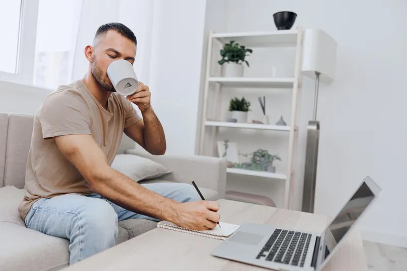 Focused man enjoying coffee while writing notes, surrounded by a modern, minimalistic workspace with plants and natural light enhancing creativity and productivity.