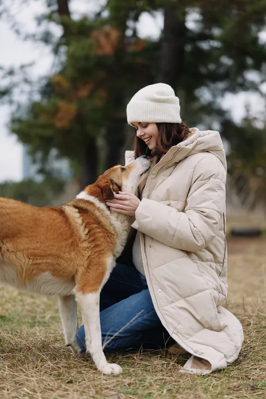 Woman kneels in a beige coat and beanie, petting a dog in an outdoor setting, sharing a calm, affectionate moment on a chilly day.