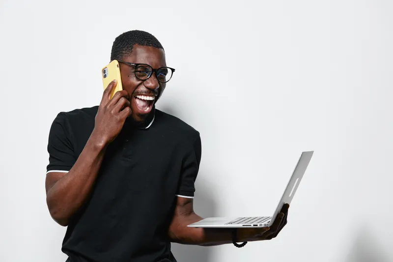 African American man with dark skin talking on phone and holding laptop, smiling and standing against white background in casual black shirt for technology and communication.