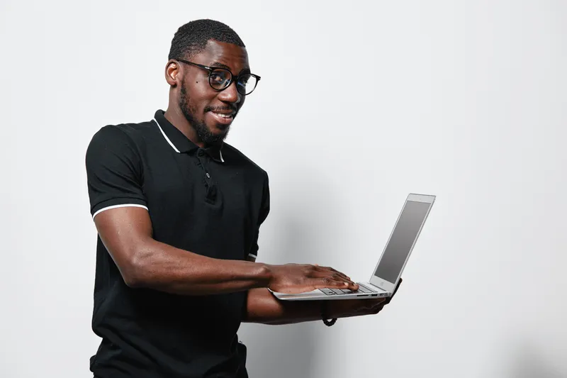 African American man with dark skin wearing glasses and black polo shirt using laptop computer for work or study on white background. Technology, casual and focused young male.