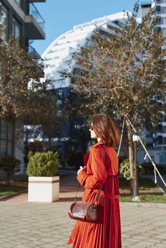 Young woman in a red dress walks through a modern urban plaza with a brown crossbody bag, capturing a candid street style moment in bright daylight.