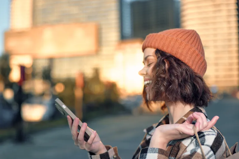 Woman with phone and smile in city wearing beanie and plaid coat, holding shopping bag during golden hour glow; candid lifestyle portrait of authenticity, mindful living and emotional storytelling.