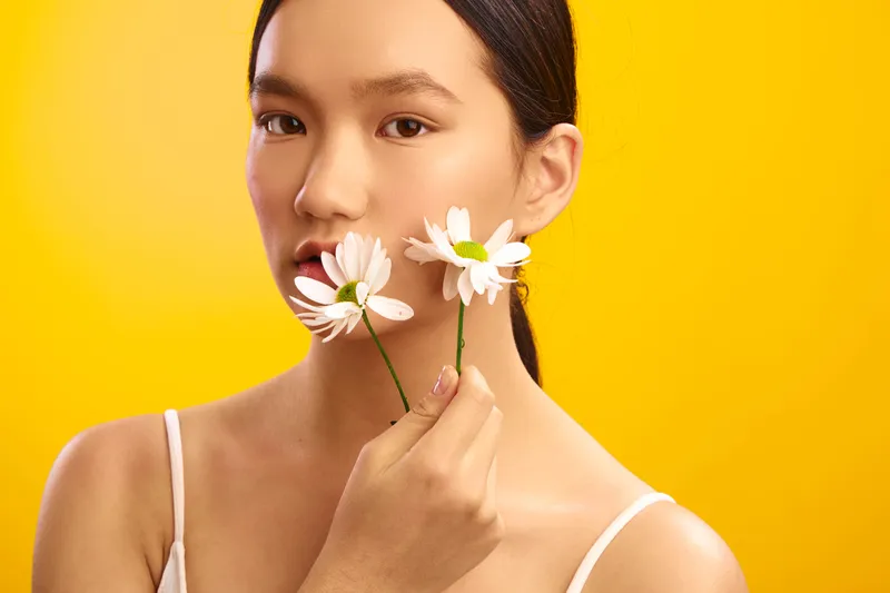 Asian woman stands against a vivid yellow backdrop, softly smiling as she holds white daisies near her lips, exuding calm confidence and graceful charm, inviting a warm, inviting mood.