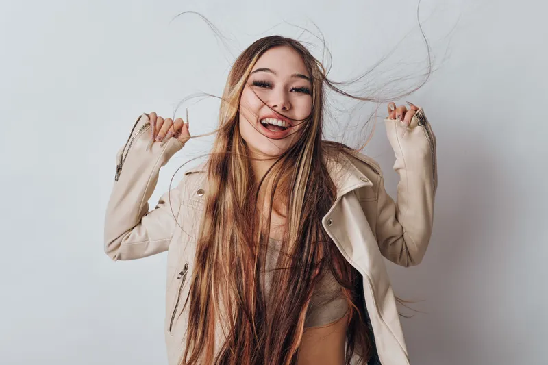Woman with long hair smiling and playing with strands in wind wearing casual jacket and casual top isolated on white background. Happy young female with joyful expression.