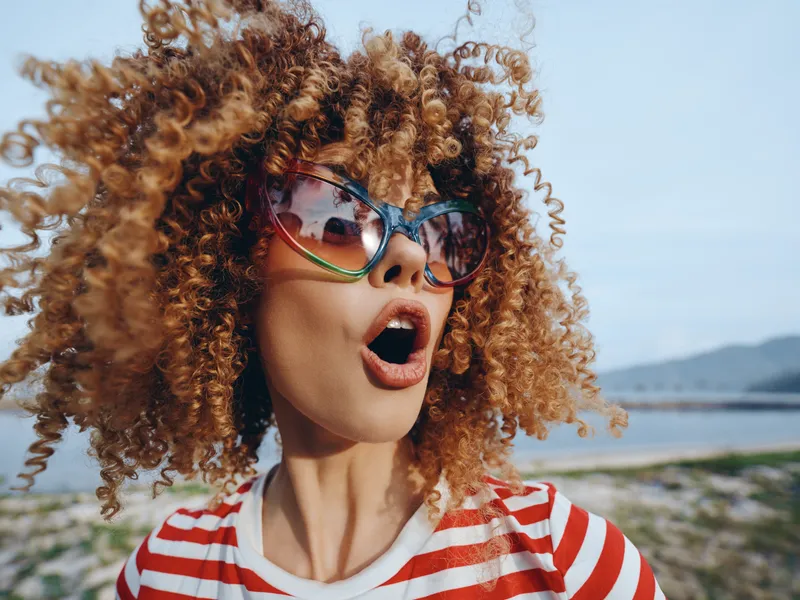 Vibrant portrait of a young woman with voluminous curly hair, oversized sunglasses, and a striped shirt, captured in a candid moment of playful surprise by the seaside.