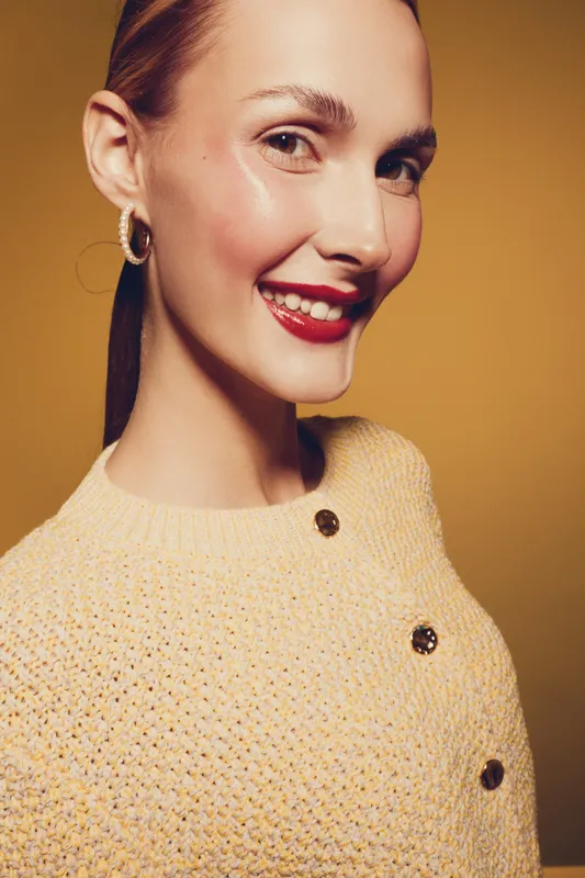 Smiling woman with red lipstick and hoop earrings wears yellow knitted sweater against warm yellow background. Portrait with natural makeup and confident expression in studio lighting.