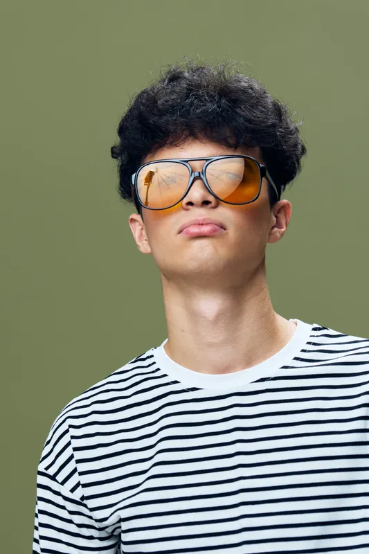 Young man with curly hair wearing large orange sunglasses and black-and-white striped shirt, posing confidently against olive green background with relaxed attitude.