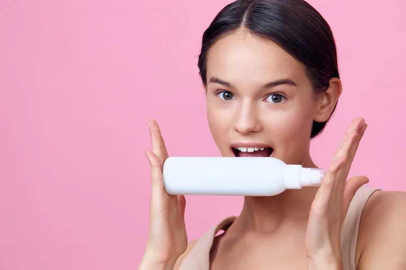 Young woman holding a white skincare product against a pink background, expressing joy and enthusiasm for beauty and skincare products, ideal for wellness marketing