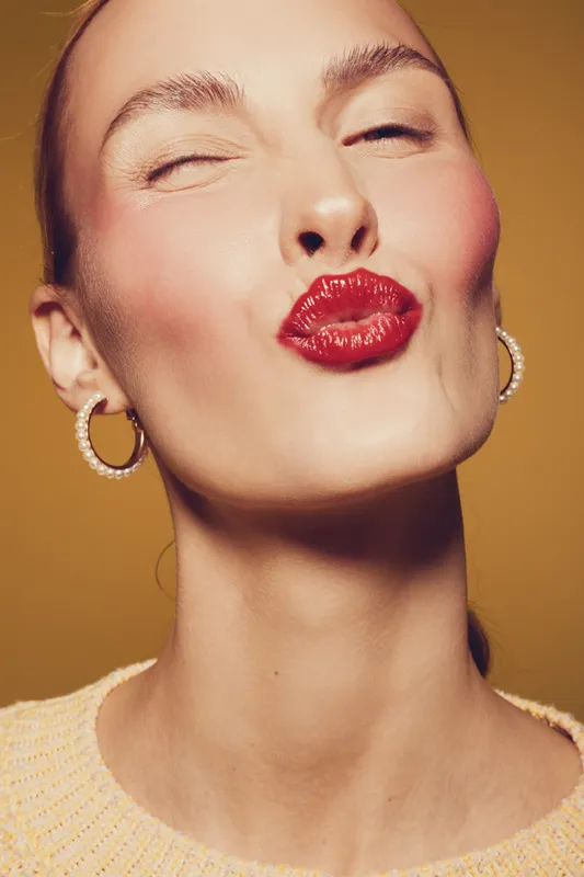 Close-up of a young woman with red lipstick making a kiss face, wearing pearl earrings and a light sweater. Studio portrait on warm brown background with soft lighting.