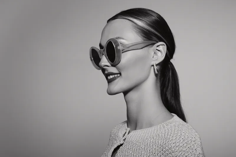 Profile of a smiling Caucasian woman with fair skin wearing large round sunglasses and a textured jacket. Studio portrait with soft lighting and minimalist background.