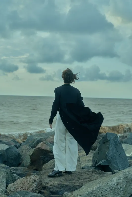 Figure standing on rocky shore in cape and tailored attire, facing the sea as wind sweeps the cape, creating a dramatic coastal moment, fusion of formalwear and rugged landscape, moody sky, expansive