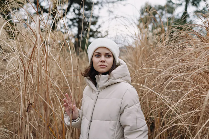 Woman in a light winter coat and white beanie stands among tall reeds in a quiet field, calm expression, soft natural light, outdoor portrait capturing chilly atmosphere and serene solitude.
