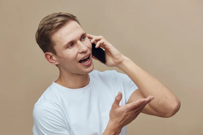 Young caucasian man talking phone excited facial expression white t-shirt casual clothes beige background digital nomad remote work concept