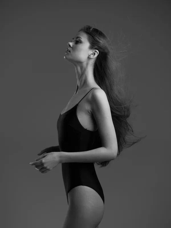 Black and white portrait of a woman in a black bodysuit with flowing hair standing sideways against gray background