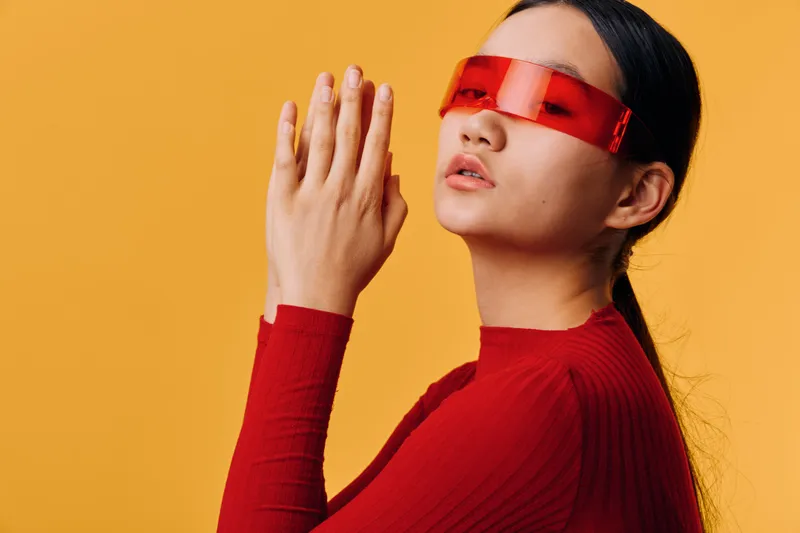 Woman in red sweater and futuristic red visor on yellow background with hands together in front of face for contemplation or meditation. Female portrait with modern style and calm expression.