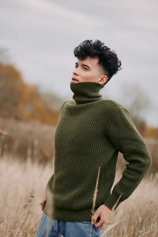 Authentic portrait of a young man in a thick olive sweater, standing in a windswept field with muted autumn tones and soft natural light. The scene conveys realism.