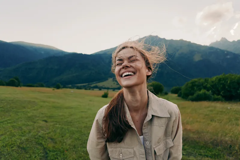 Cheerful woman outdoors, smiling with eyes closed, sunlit moment in a field near mountains, casual jacket, light breeze, natural colors, lush greenery, carefree mood today