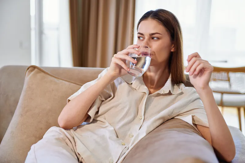 Woman on sofa drinking water from a glass at home, relaxed portrait on couch in casual loungewear, hydration and wellness moment, cozy interior with natural light and calm expression