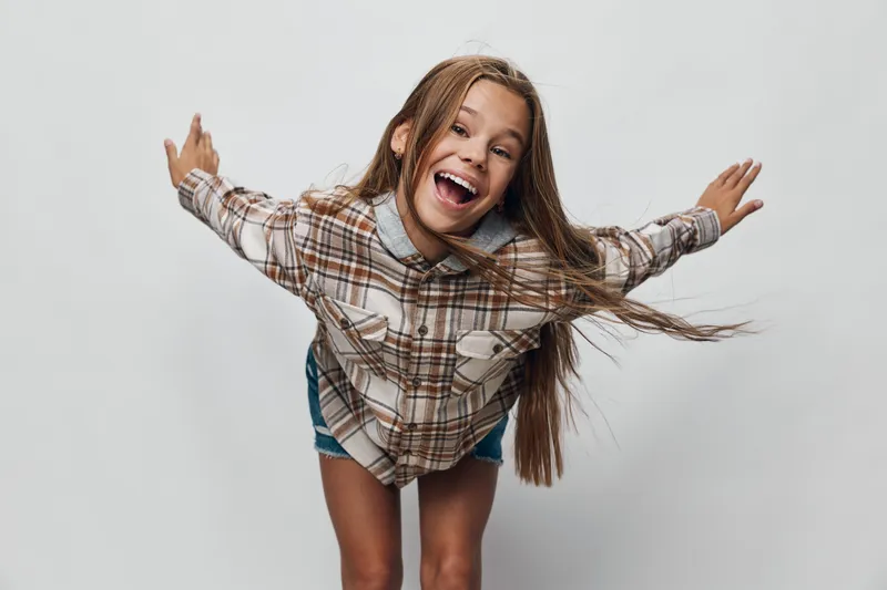 Girl smiling and leaning forward with arms outstretched wearing plaid shirt and shorts against plain background. Cheerful child with long hair and happy expression in studio.