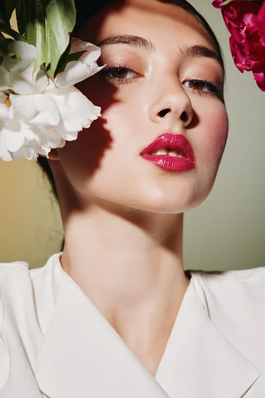 Close-up of a young woman with glossy red lips and flawless skin framed by white and red flowers. Elegant beauty fashion concept with creative lighting and shadows.