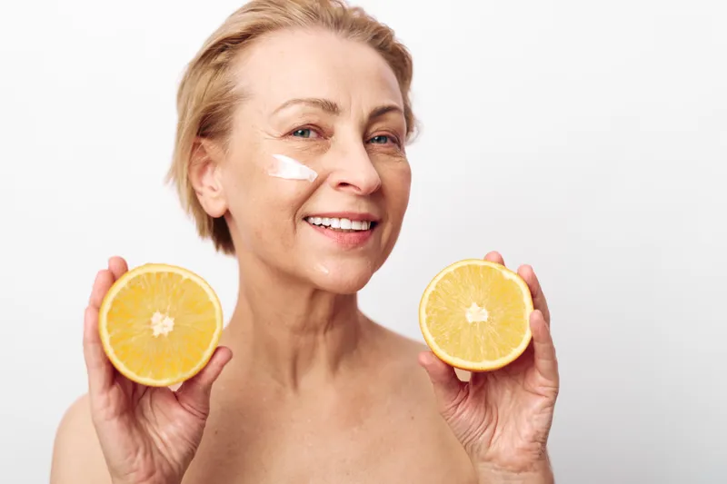 Smiling mature woman with short hair holds two orange halves near face with white cream on cheek, isolated on white background. Concept of natural skincare, healthy lifestyle, and freshness.