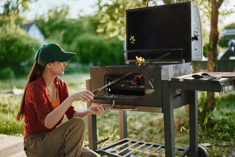 A young woman grilling outdoors, wearing a green cap and red shirt, displaying joy and focus while preparing food in a summer garden
