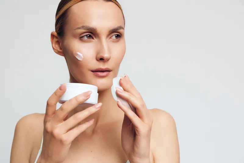 Young woman with clean skin applying white face cream on cheek using cotton pad. Skincare routine concept with soft natural light and neutral background for beauty and wellness.