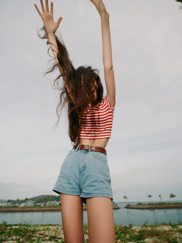 Young woman stands outdoors with arms raised high, carefree pose wearing a striped crop top and denim shorts, bright sky, and a serene waterline in the distance.